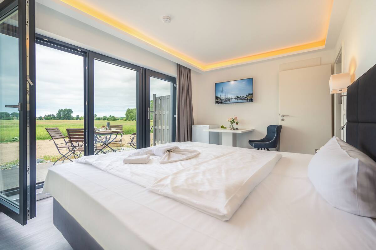 Een moderne hotelkamer met een groot bed, een bureau en een stoel. Er is uitzicht op het omliggende landschap door de glazen deuren.