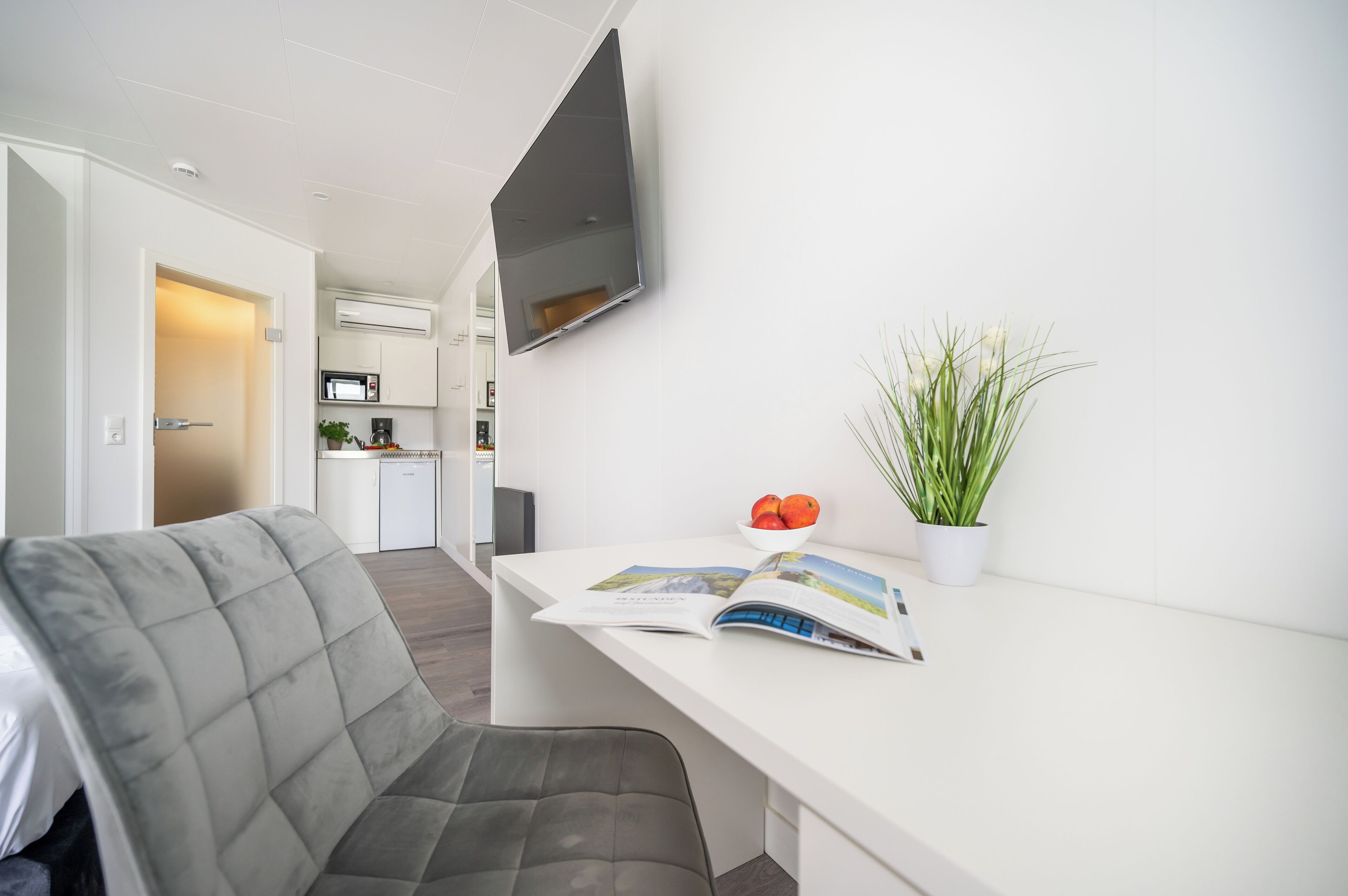 Habitación moderna y luminosa con una butaca tapizada en gris, un escritorio blanco con una revista, una planta en maceta y un televisor de pantalla plana montado en la pared.