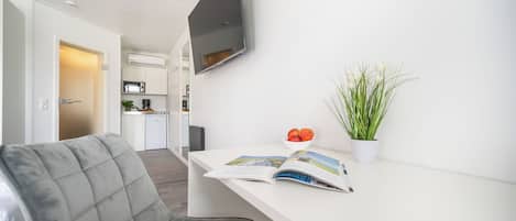 Habitación moderna y luminosa con una butaca tapizada en gris, un escritorio blanco con una revista, una planta en maceta y un televisor de pantalla plana montado en la pared.