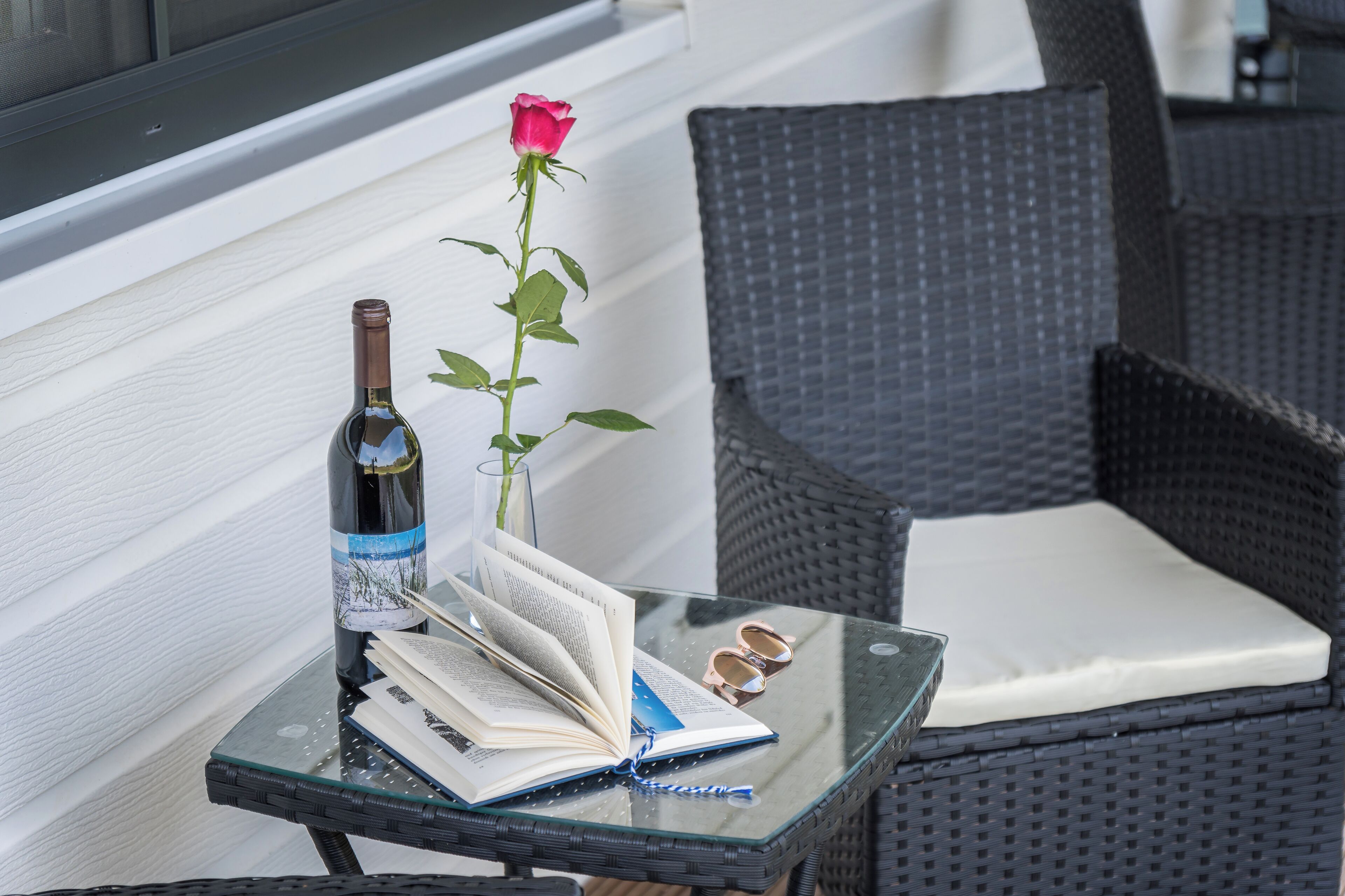 Une table en verre sur laquelle se trouvent une bouteille de vin, un livre ouvert, une paire de lunettes et une rose.