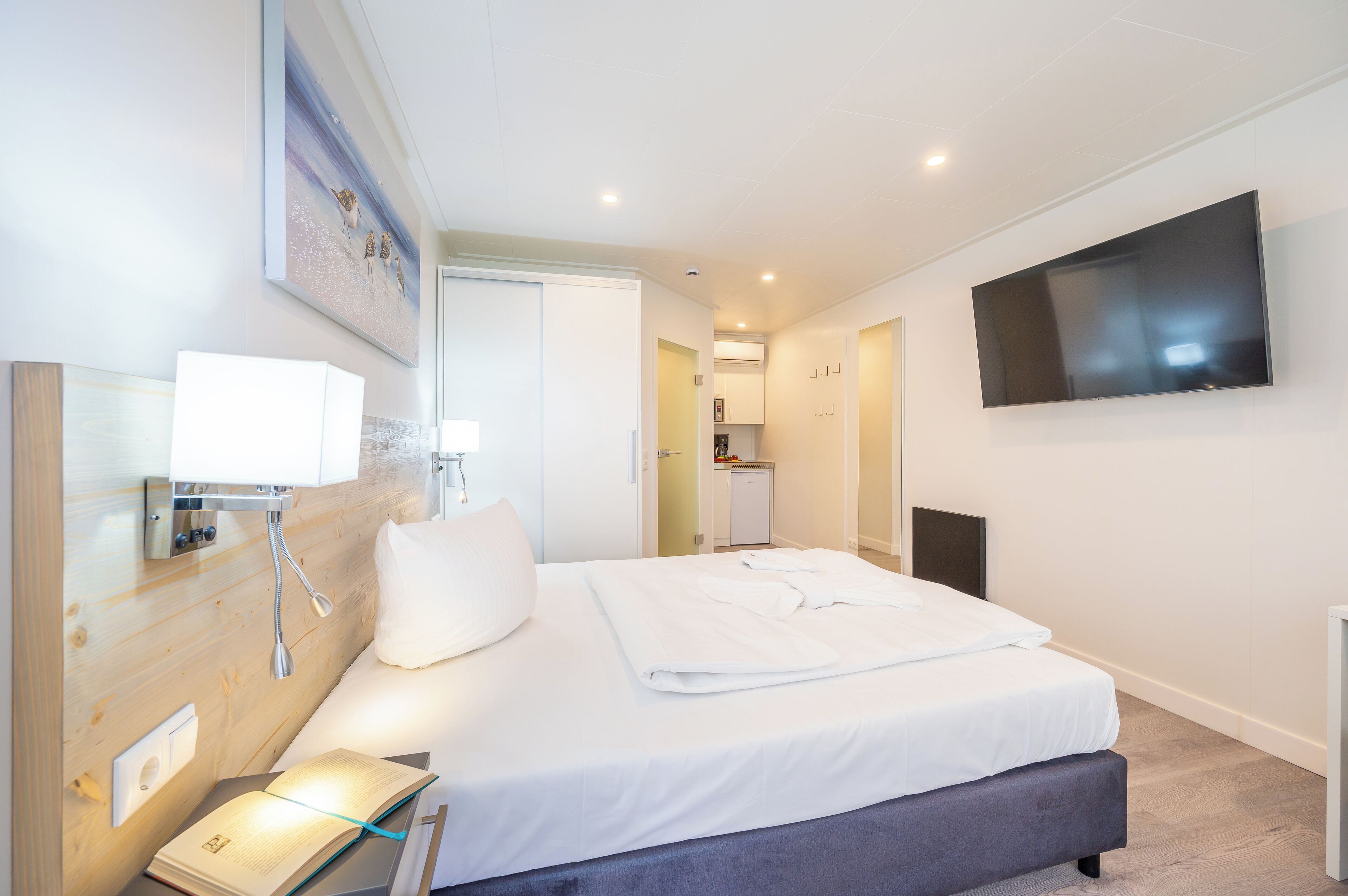 A modern hotel room with a large bed, a flat-screen TV mounted on the wall, a bedside table with a lamp, and a small kitchenette in the background.