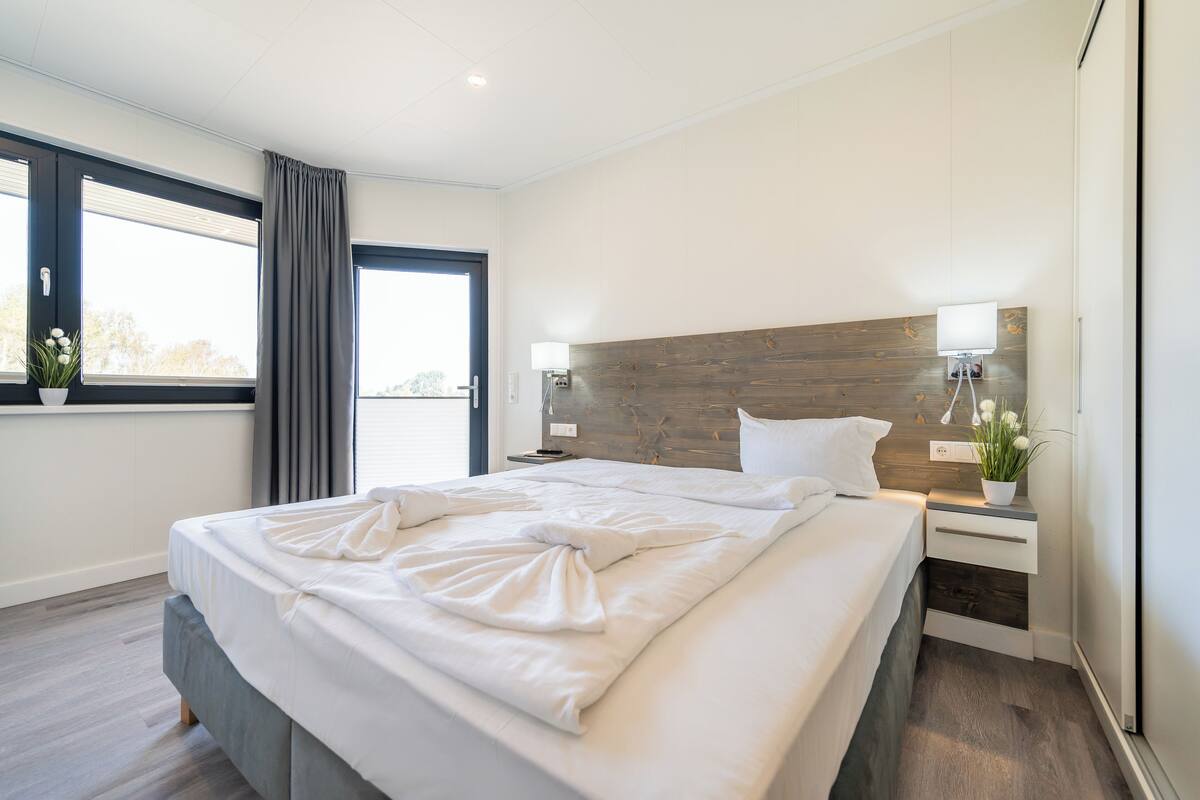 Een moderne slaapkamer met een groot bed, nachtkastjes, een houten hoofdbord en een raam met gordijnen.