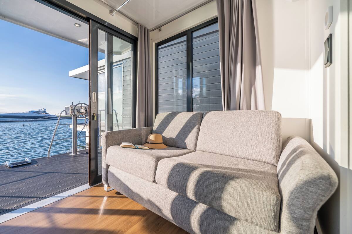 Een modern interieur met een bank, een hoed en een boek op een terras met uitzicht op een waterlichaam.