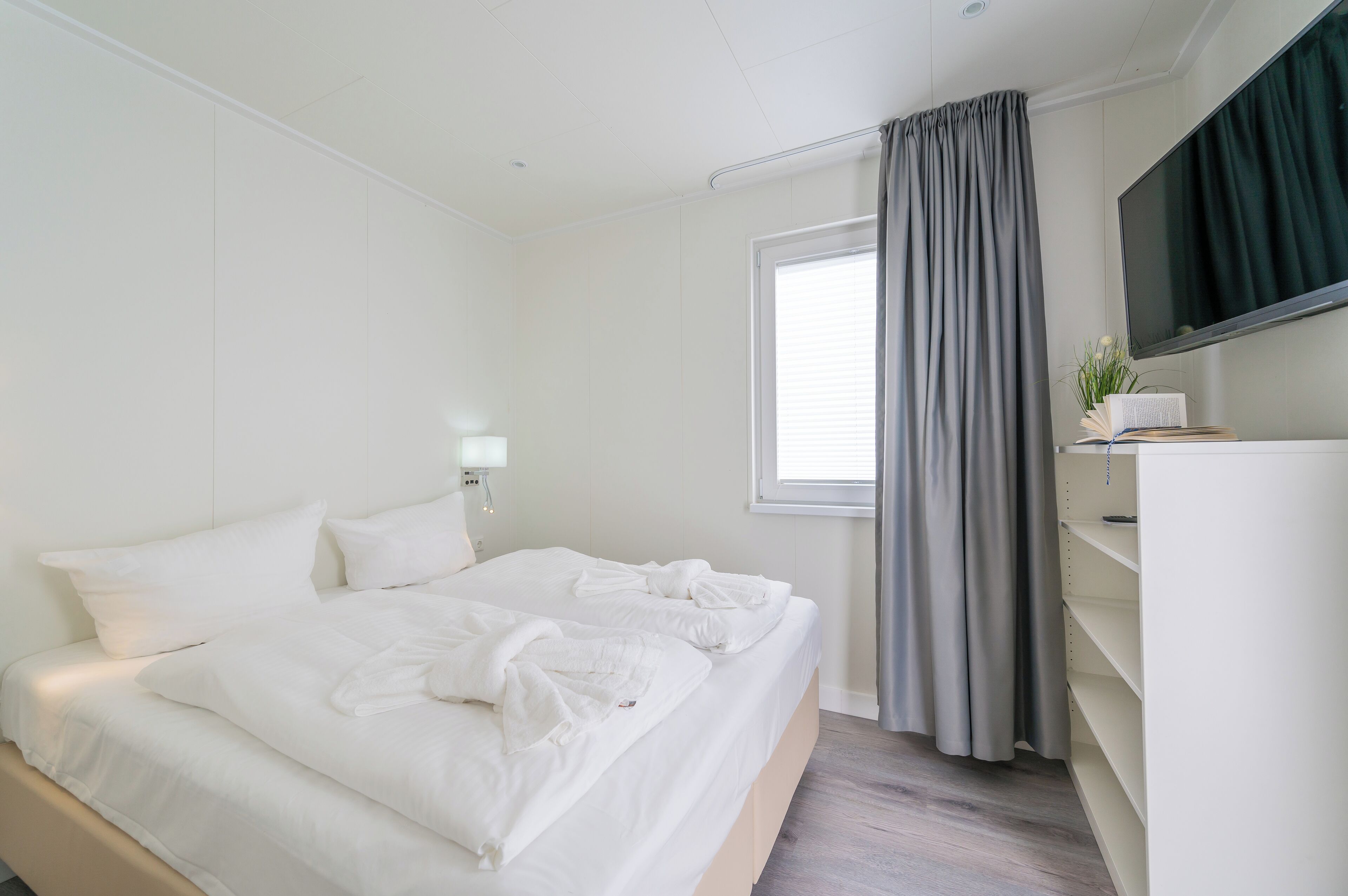 A modern hotel room with a large bed, a flat-screen TV, a bedside table with a lamp, and a window with curtains.