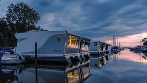 Une rangée de camping-cars est amarrée dans une marina, sur une eau calme qui reflète les bateaux, avec un coucher de soleil en arrière-plan.