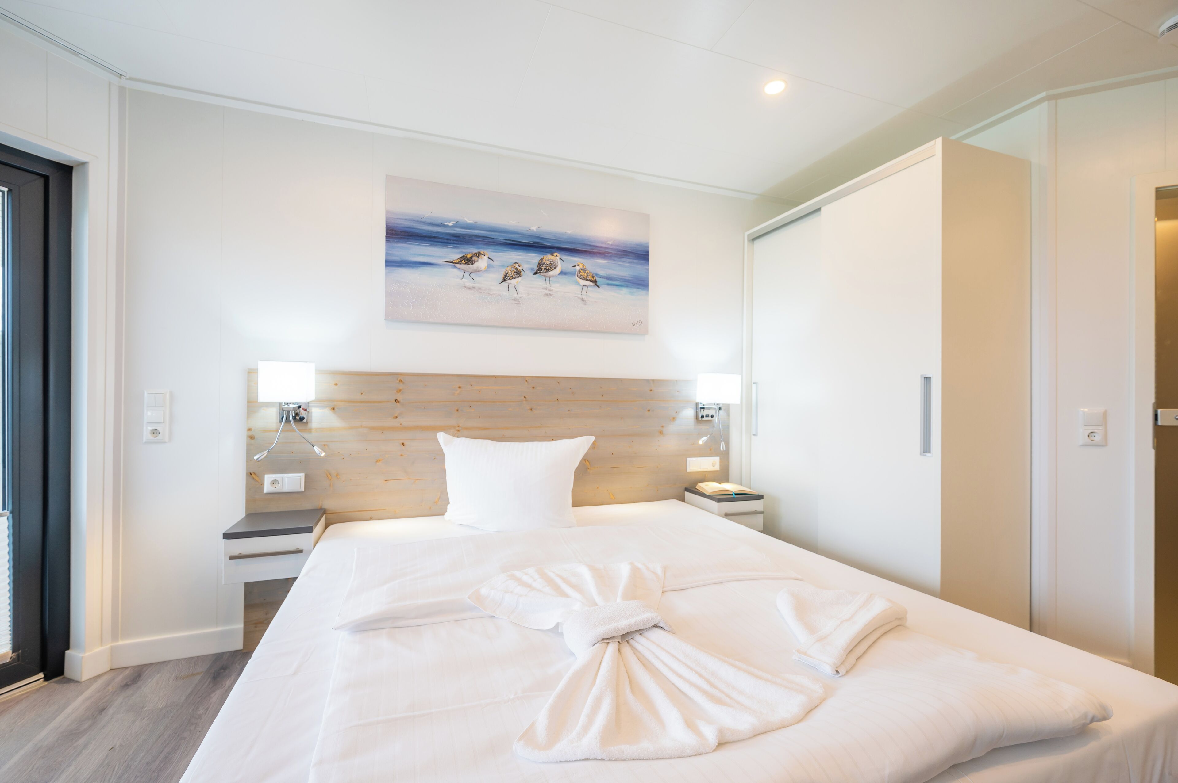 Une chambre d’hôtel avec un grand lit, une tête de lit en bois, un tableau représentant des oiseaux accroché au mur et un lit parfaitement fait, agrémenté de serviettes.