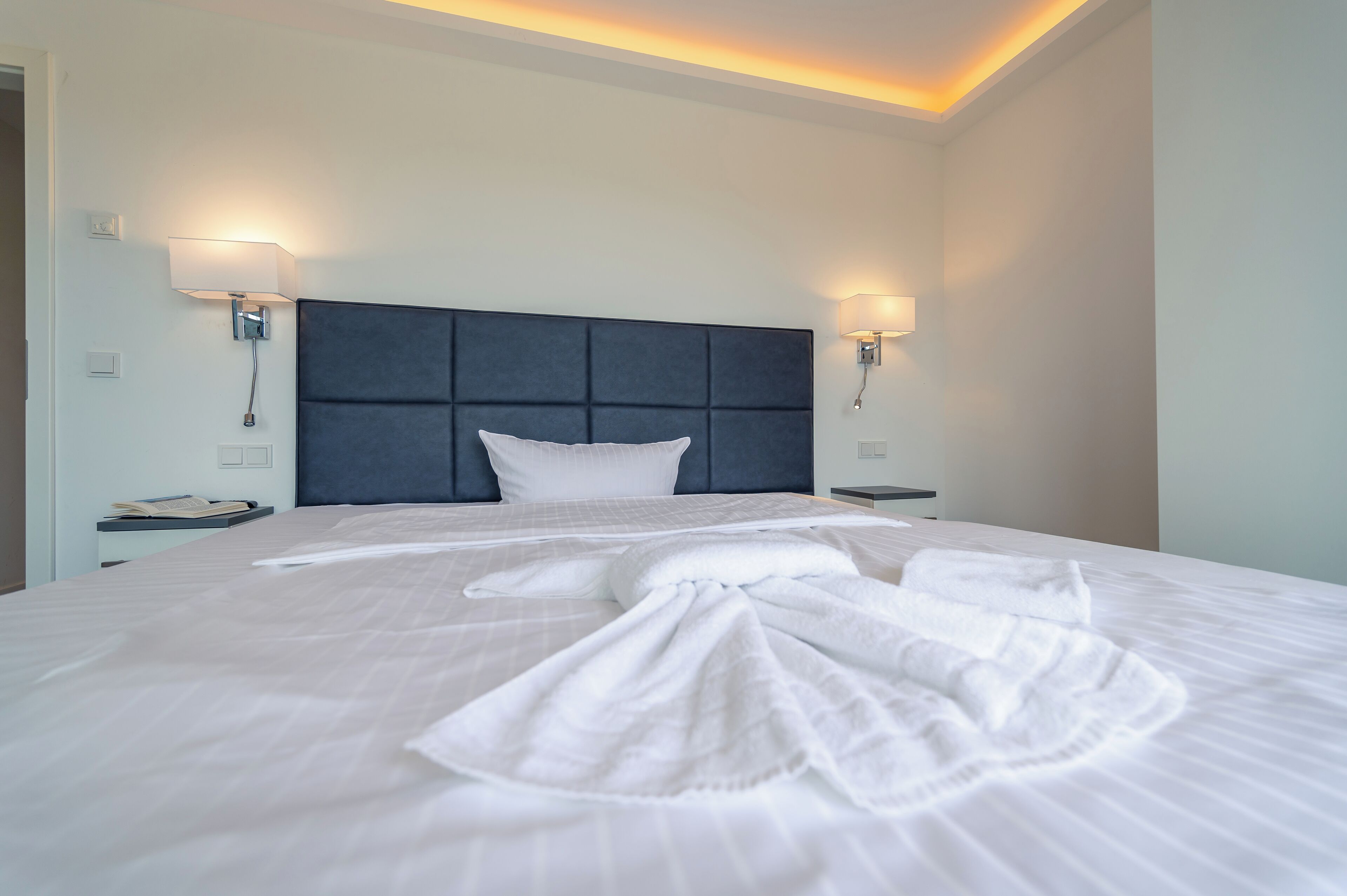Una cama bien hecha con un cabecero oscuro, dos lámparas de noche y una mesita de noche con un teléfono.
