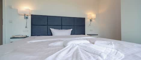 Una cama bien hecha con un cabecero oscuro, dos lámparas de noche y una mesita de noche con un teléfono.