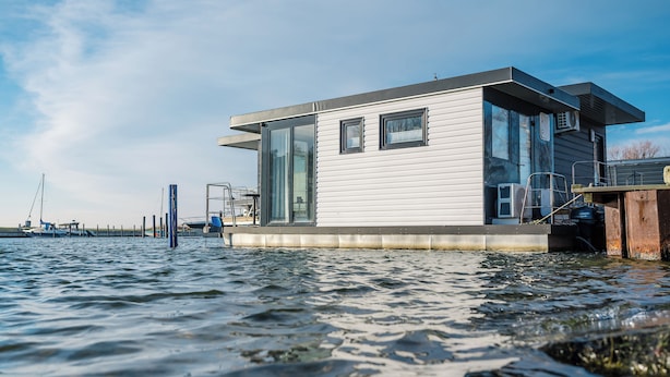 Une maison flottante moderne, dotée de grandes fenêtres et d’un toit plat, située sur une étendue d’eau calme.
