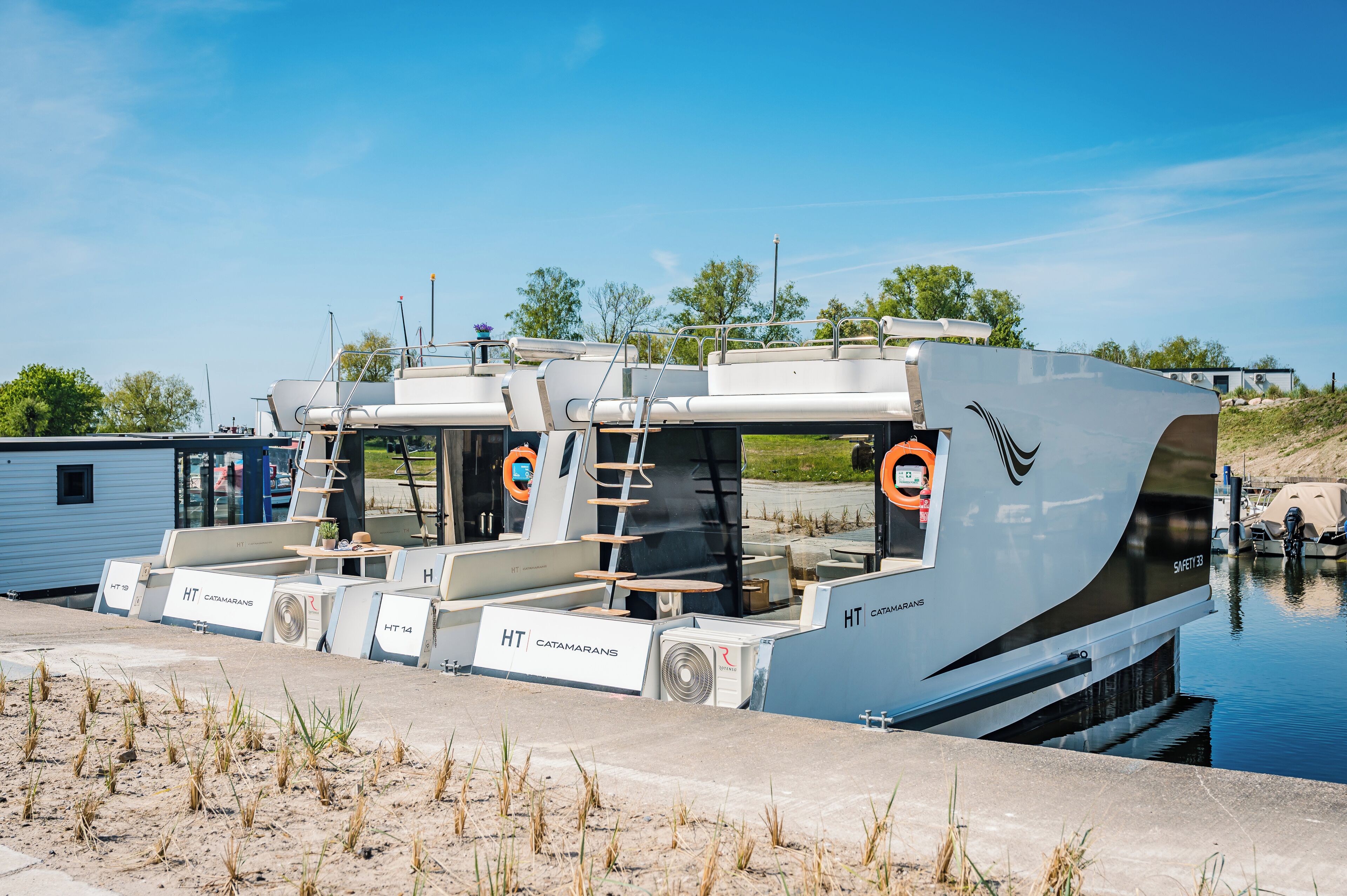 A houseboat with multiple decks, labeled "HT Catamarans," docked at a canal.