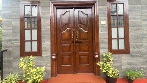 A double wooden door with glass panels, flanked by two windows with wooden frames and glass panes, leading to a red-carpeted entrance. - Villa Mercara (Madikeri)