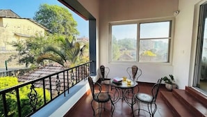 A balcony with a table and chairs, a view of greenery and buildings, and a window with a view of the landscape. - Villa Mercara (Madikeri)