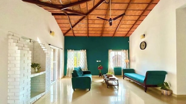 A spacious living room with a high ceiling, a brick fireplace, two green sofas, a coffee table, and a ceiling fan. - Villa Mercara (Madikeri)