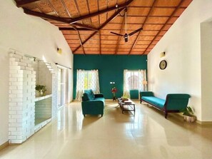 A spacious living room with a high ceiling, a brick fireplace, two green sofas, a coffee table, and a ceiling fan. - Villa Mercara (Madikeri)