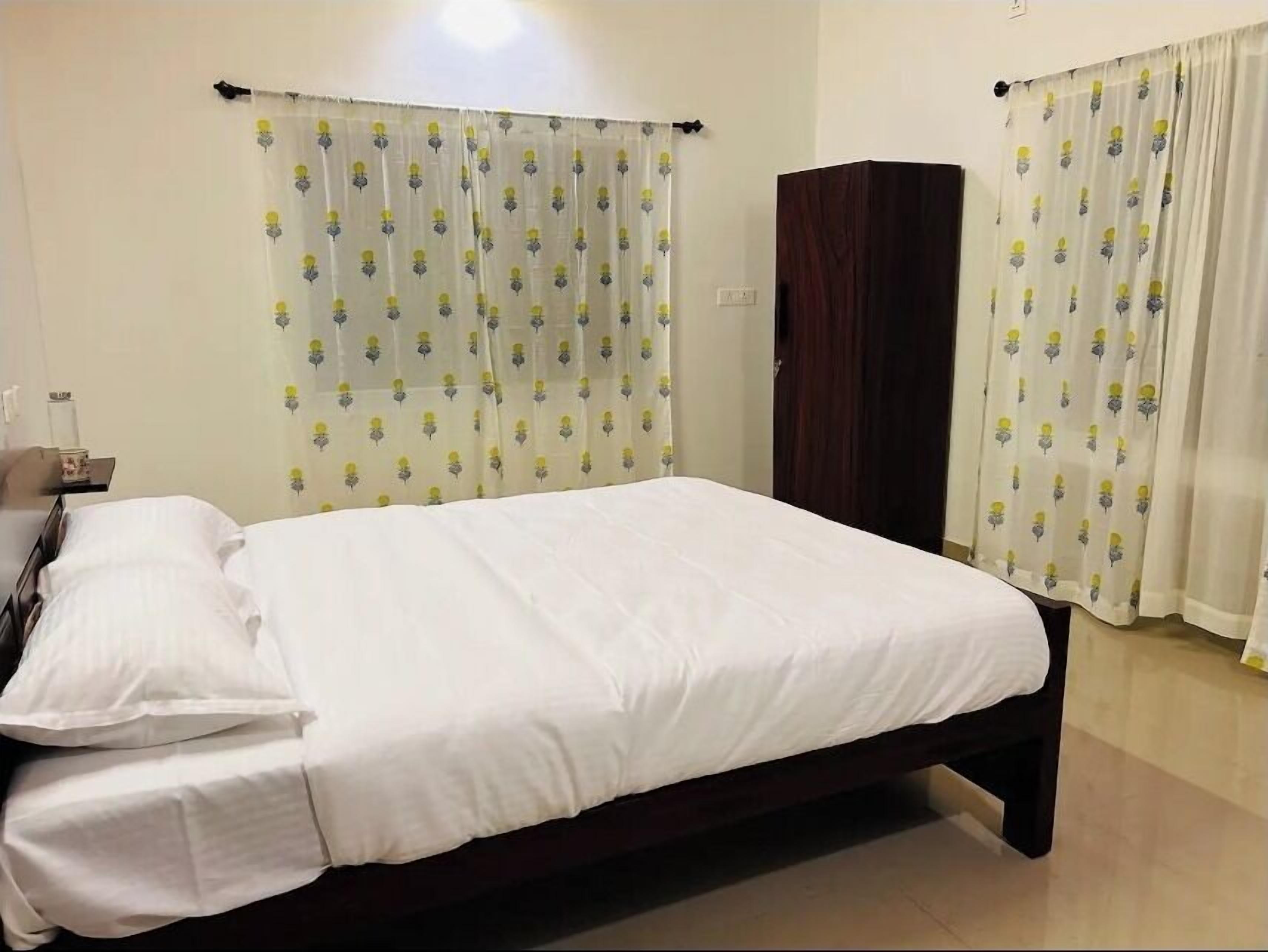 A neatly made bed with white linens in a room with a wooden wardrobe and a curtain with a floral pattern.