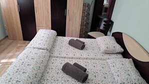 A neatly made bed with floral bedding, two rolled towels, and a small bedside table with a chair.