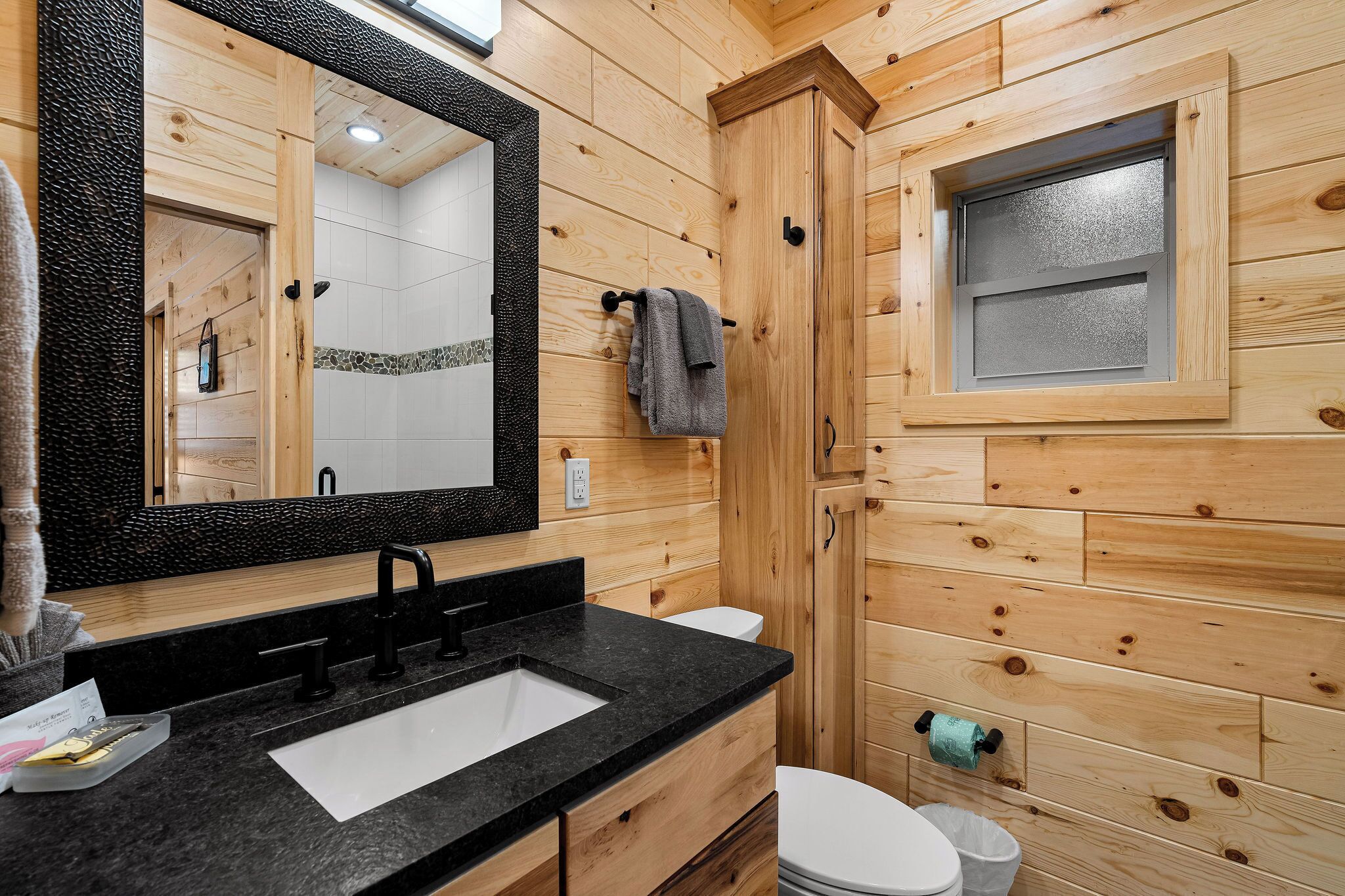 A wooden cabin bathroom with a black sink, a mirror, a towel rack, and a window.