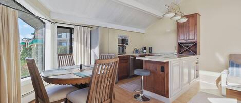 A modern kitchen with a central island, a dining area with a table and chairs, and a view through large windows.