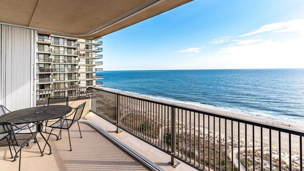 A balcony with a table and chairs, overlooking a beach and a high-rise building. - HPN-Stunning 2 Bedroom Direct Ocean Front Condo (Ocean City)
