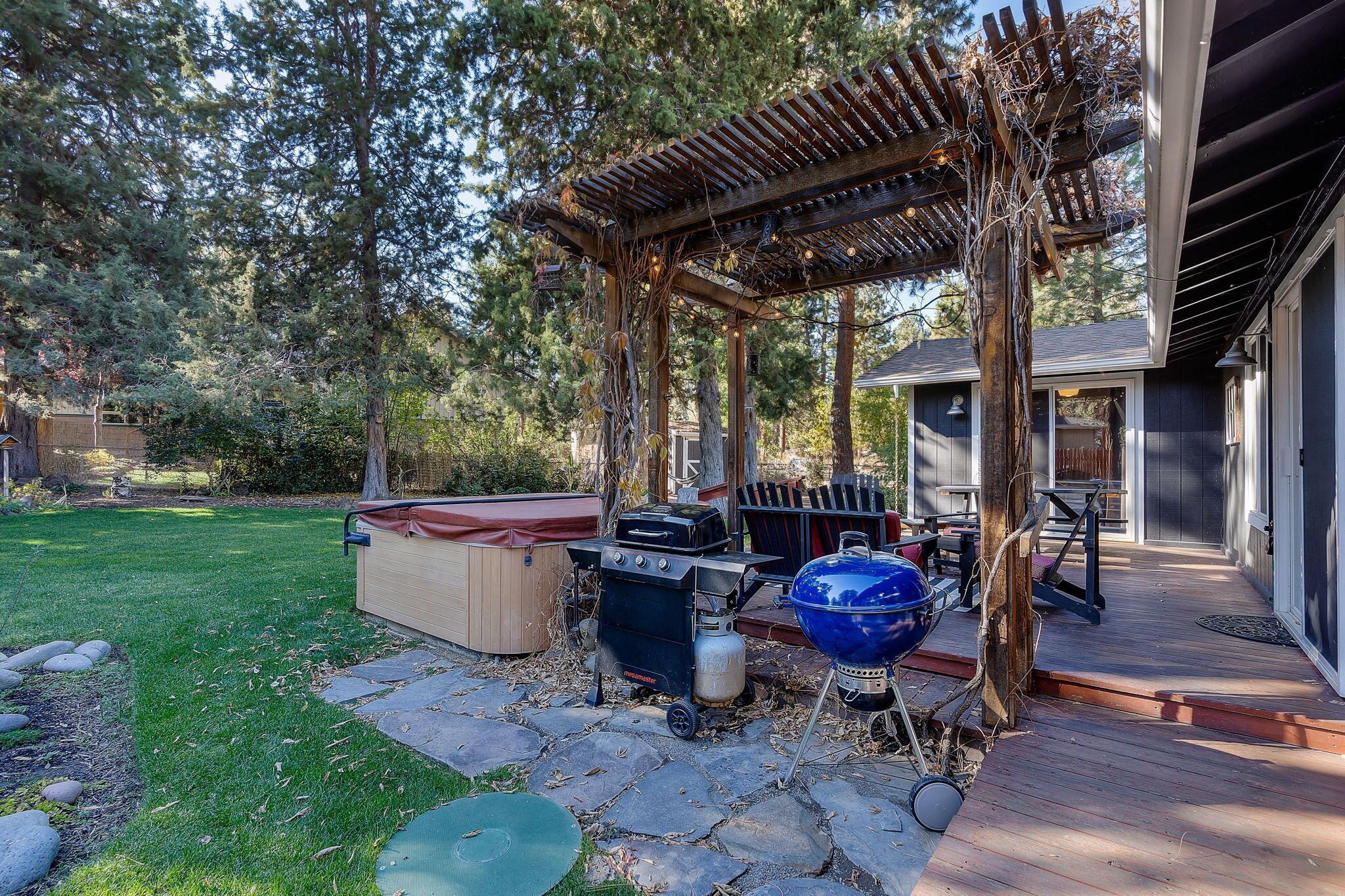 Ein Garten mit einer Holz-Pergola, einem Grill, einem Whirlpool und einer Terrasse, die zu einem Haus führt.