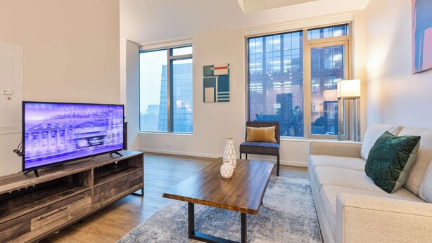 Un salon moderne doté d’un téléviseur à écran plat, d’un meuble de rangement en bois, d’une table basse, d’un canapé et de grandes fenêtres offrant une vue sur la ville.