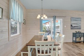 A dining area with a wooden table and white chairs, a chandelier, and a view through a door to the outside.