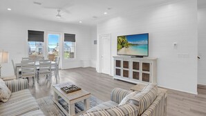 A spacious living room with a dining area, white walls, and a large TV on a cabinet. - West Side Cottages CC by Liquid Life (Gulf Shores)