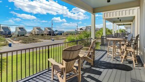 A covered porch with wooden chairs and a table, overlooking a street with houses and a clear sky. - West Side Cottages CC by Liquid Life (Gulf Shores)