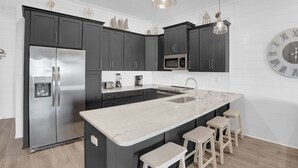 A modern kitchen with dark cabinets, a stainless steel refrigerator, a marble countertop, and a large wall clock. - West Side Cottages CC by Liquid Life (Gulf Shores)