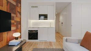 Una cocina moderna con armarios blancos, microondas integrado y horno. Se ve un televisor de pantalla plana en la pared y una mesita con libros y una lámpara.