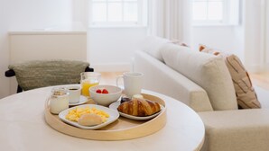 En bakke med croissanter, røræg og et glas appelsinjuice på et rundt bord i et lyst rum med en hvid sofa og et vindue.