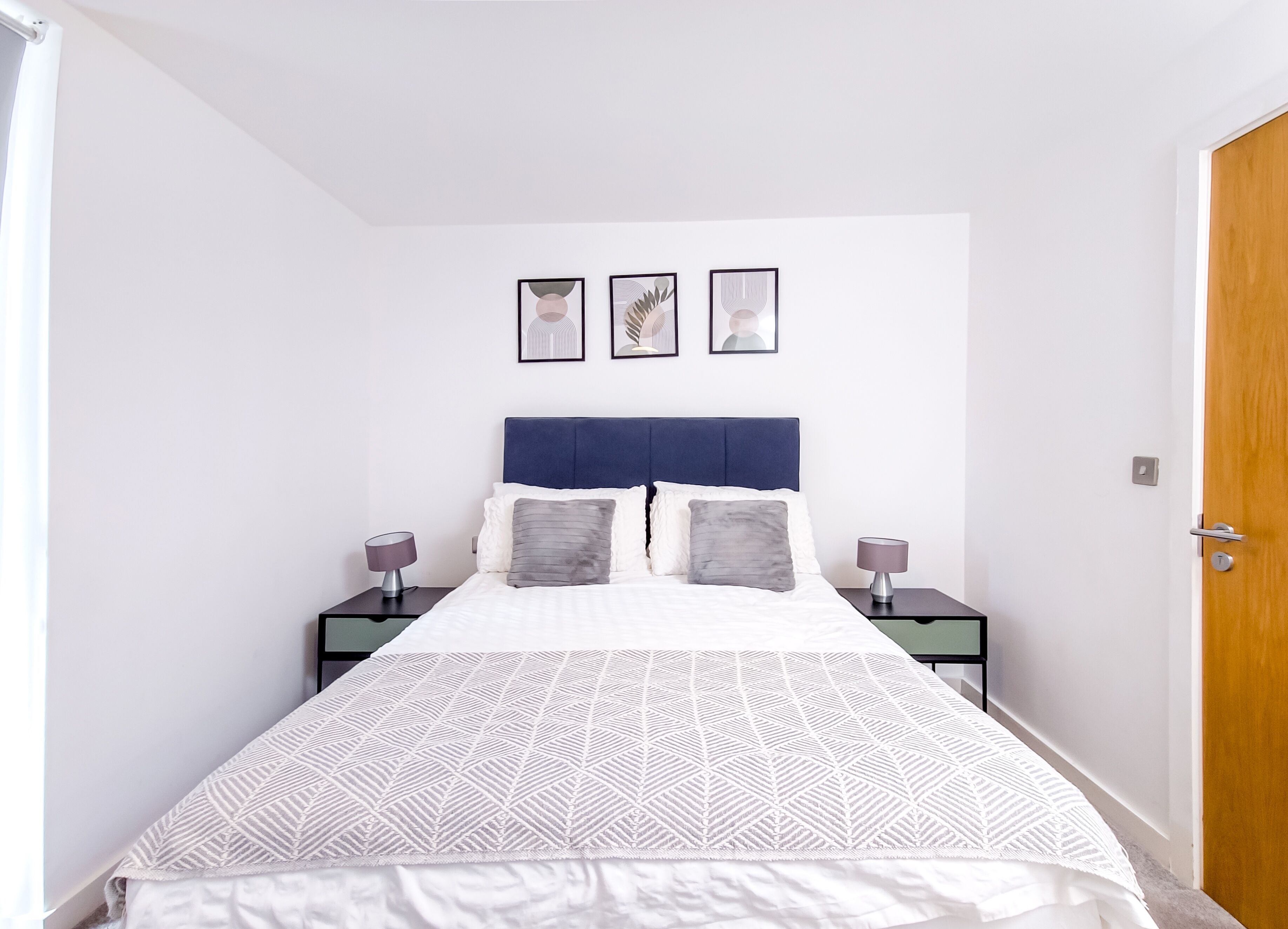 A bedroom with a bed, two nightstands, a wooden door, and three framed pictures on the wall.