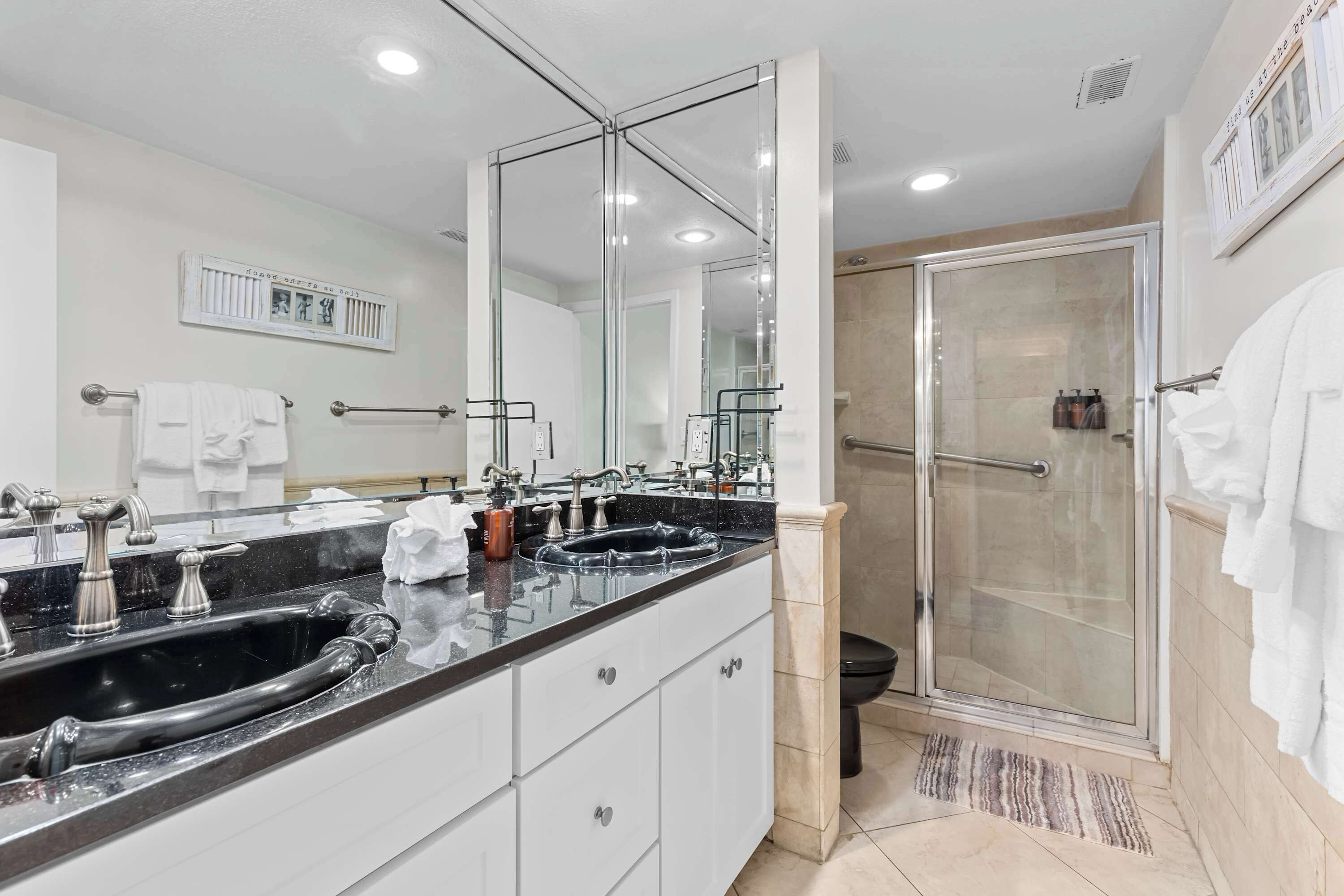 Un baño moderno con un espejo grande, un lavabo negro, armarios blancos y una ducha con mampara de vidrio.
