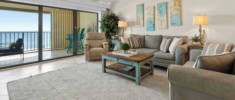 A spacious living room with a large window, a ceiling fan, a sofa set, a coffee table, and a view of the ocean.