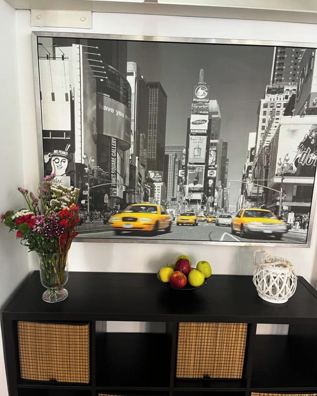 Une photographie en noir et blanc représentant un paysage urbain, accrochée au mur, un vase de fleurs, un bol de pommes et un panier tressé, le tout posé sur une console noire.