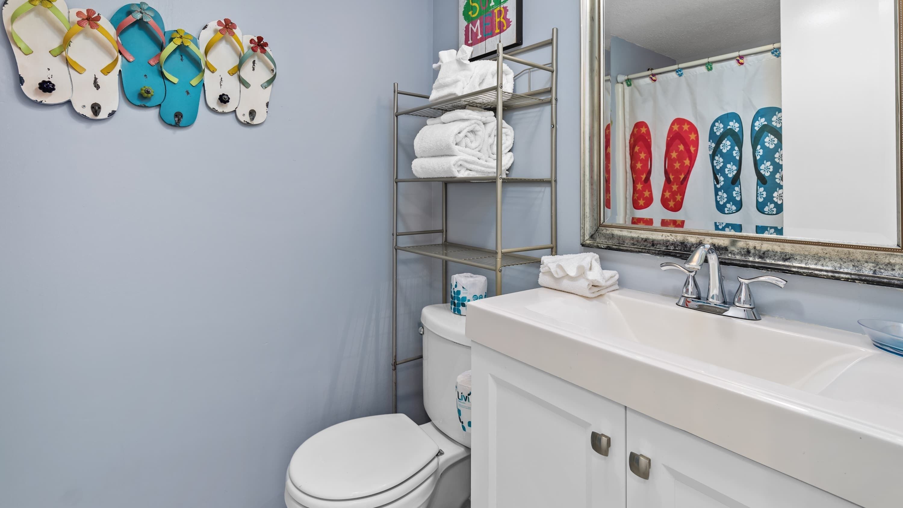 A bathroom with a white sink, a mirror, a toilet, and a shelf with towels and flip-flops.