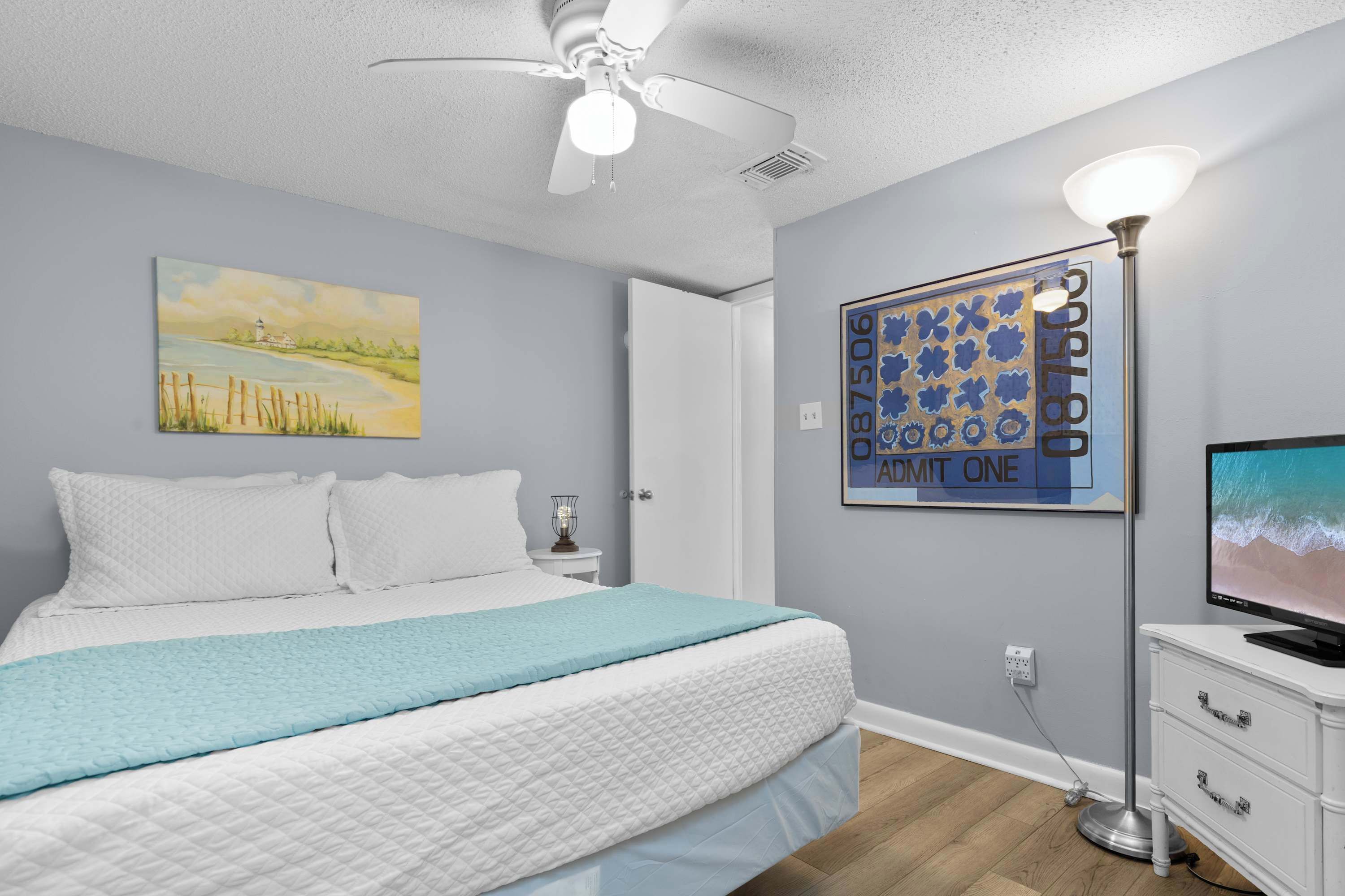 A bedroom with a bed, a ceiling fan, a painting, a lamp, and a television.