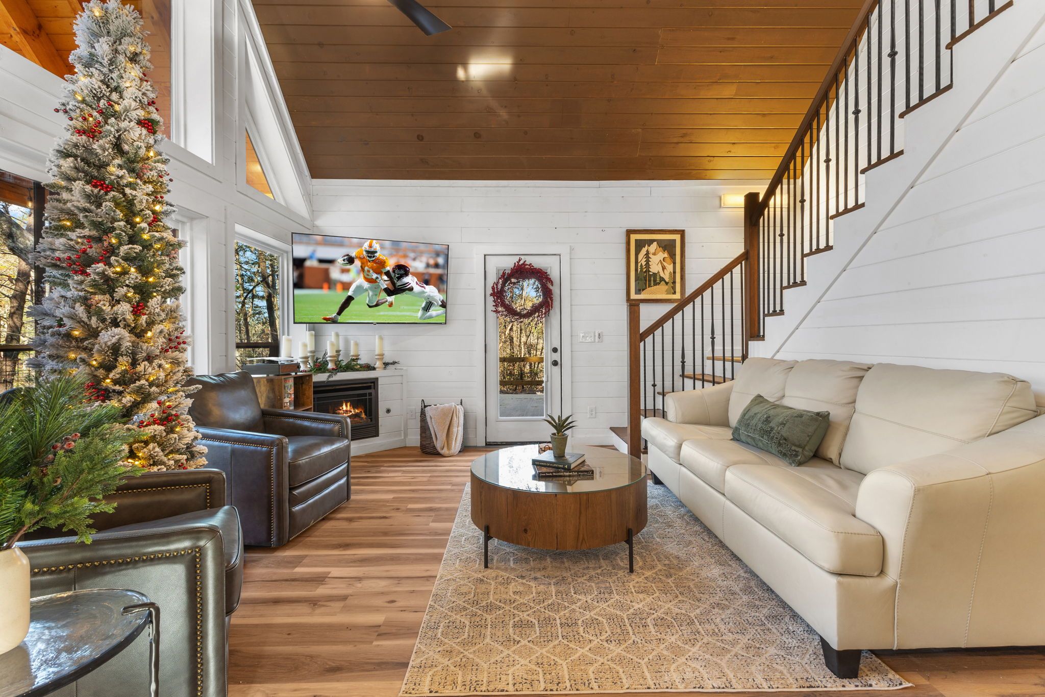 A cozy living room with a Christmas tree, leather sofas, a coffee table, and a TV displaying a football game.