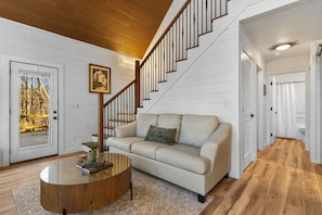 A living room with a beige sofa, a glass coffee table, a wooden staircase, and a white door leading outside. - Arcade | Private Hot Tub | Firepit | Fireplace | Close to Attractions (Sevierville)