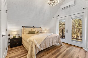 Een slaapkamer met een bed, nachtkastjes, een kroonluchter en grote ramen met uitzicht op bomen.