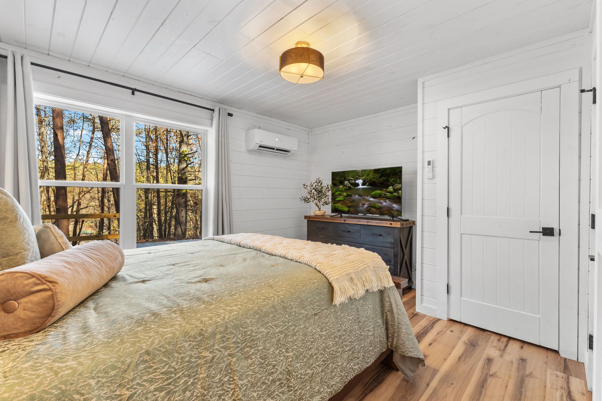 Een slaapkamer met een groot bed, een televisie op een dressoir, een raam met uitzicht op bomen en een witte deur.