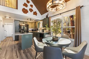 A modern kitchen with a central island, dining area with chairs, and a large window with a view of trees. - Arcade | Private Hot Tub | Firepit | Fireplace | Close to Attractions (Sevierville)