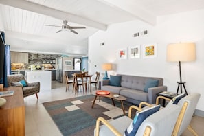 Uma sala de estar moderna com sofá, poltronas, mesa de centro e área de jantar com mesa e cadeiras. Uma cozinha ao fundo, com geladeira e fogão.