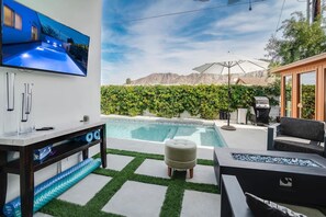 A backyard with a swimming pool, a TV mounted on the wall, outdoor furniture, and a hedge. - The Elemental Retreat- Pool Spa and Sauna (La Quinta)