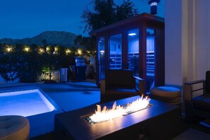 A pool area with a fire feature, seating, and a covered structure with lights. - The Elemental Retreat- Pool Spa and Sauna (La Quinta)