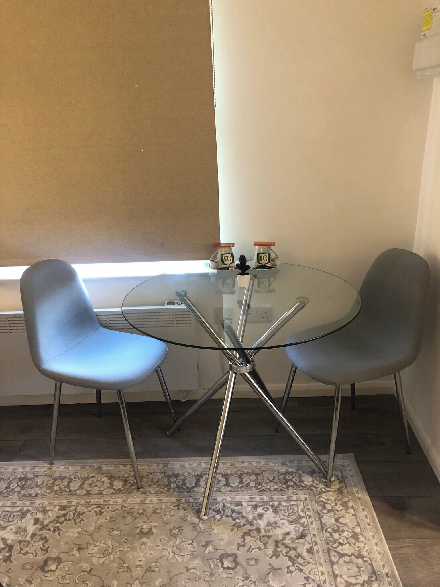 A small, modern room with a glass table surrounded by two chairs, a radiator, and a patterned rug.
