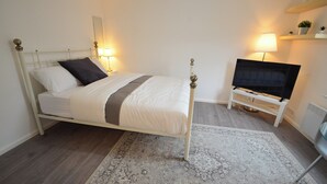 A neatly arranged bedroom with a bed, bedside lamps, a television on a stand, and a small shelf with plants. - Hackney E5 - Perfect for Couples Getaway (London)