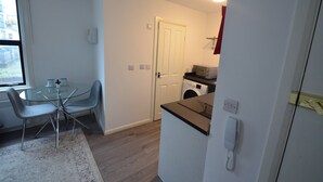A compact kitchen with a washing machine, a dining area with a glass table and chairs, and a telephone on the wall. - Hackney E5 - Perfect for Couples Getaway (London)