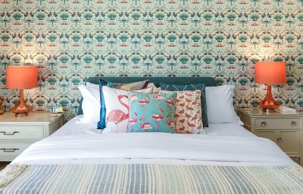 A neatly made bed with multiple pillows, a patterned wallpaper, and two bedside lamps.