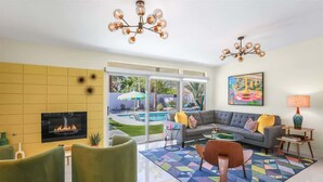 Un salon moderne avec une cheminée, des fauteuils verts, un canapé avec des coussins, un tapis coloré et une grande fenêtre donnant sur une piscine et un jardin.